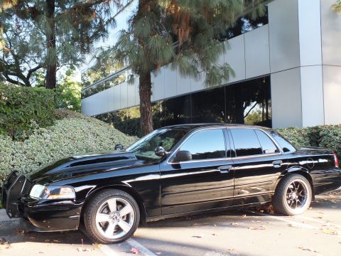 2003 Ford Crown Victoria - Driver-side view of a black full-size sedan, front grille guard visible, sitting low on dark alloy wheels.
