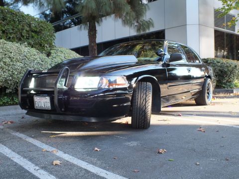 2003 Ford Crown Victoria - Ground-level front-quarter of a lowered black sedan, grille guard and EVIL VIC plate, wheel filling the arch.