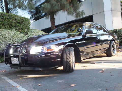 2003 Ford Crown Victoria - Wide low-angle front-quarter of a glossy black sedan, push bar up front and EVIL VIC plate visible.