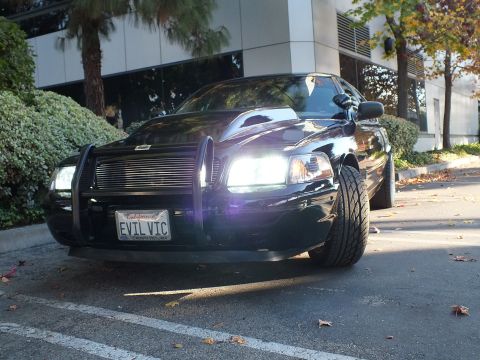 2003 Ford Crown Victoria - Close low front-quarter of a black sedan, lit headlights, push bar framing an EVIL VIC plate.