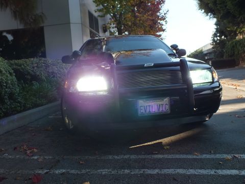 2003 Ford Crown Victoria - Head-on of a black sedan with headlights flared bright, push bar and billet grille over EVIL VIC plate.