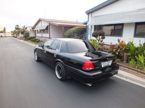 2003 Ford Crown Victoria - Rear three-quarter of a black sedan with dual tailpipes and dark wheels parked on a residential drive.