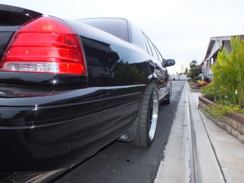 2003 Ford Crown Victoria - Low rear-quarter view of a black Crown Victoria sedan with red taillight and a polished-lip wheel near the curb.