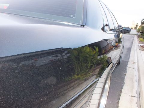 2003 Ford Crown Victoria - Reflective black sedan rear quarter and side mirror catching foliage, polished wheel lip at lower edge.