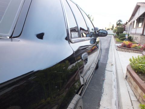 2003 Ford Crown Victoria - Black sedan front door, mirror and window-decal sail panel parked along a curb beside a planted bed.