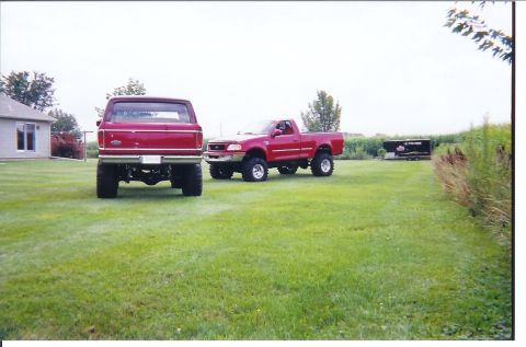 1978 Ford Bronco - Scanned print of two lifted red Ford 4x4s parked on a mowed yard beside a cornfield: a full-size Bronco shown from the rear with its tailga…