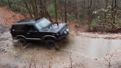1986 Ford Bronco - A black SUV navigates a muddy obstacle on a wooded trail.