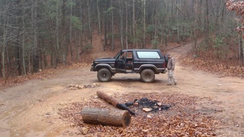 1986 Ford Bronco - A black Ford Bronco is parked on a dirt path surrounded by trees, with a person standing near the rear of the vehicle.
