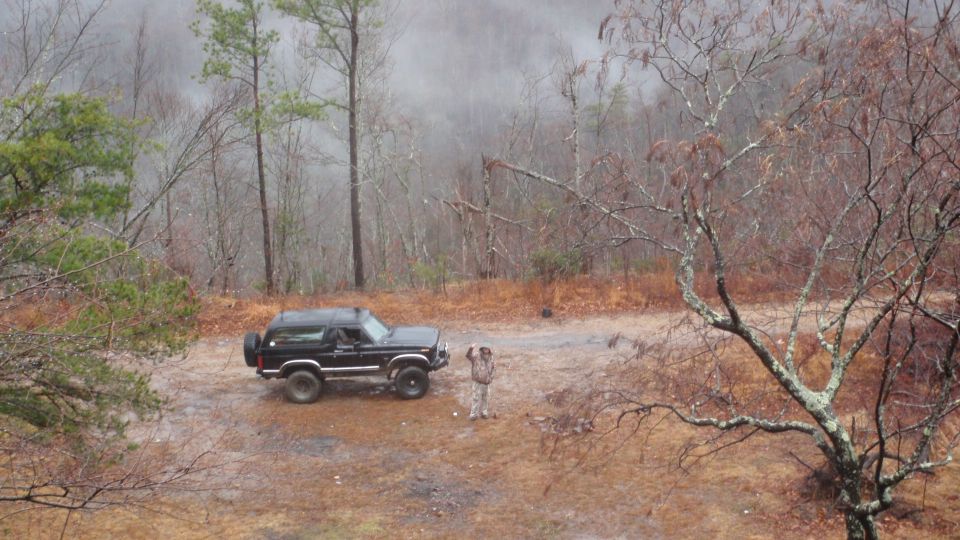 Black Ford Bronco parked on dirt road, wooded area, light snow falling. Person standing near vehicle