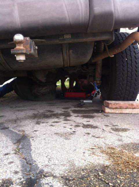 1989 Ford Bronco - A person works on the underside of a vehicle, with the fuel tank, hitch, and a tire clearly visible.