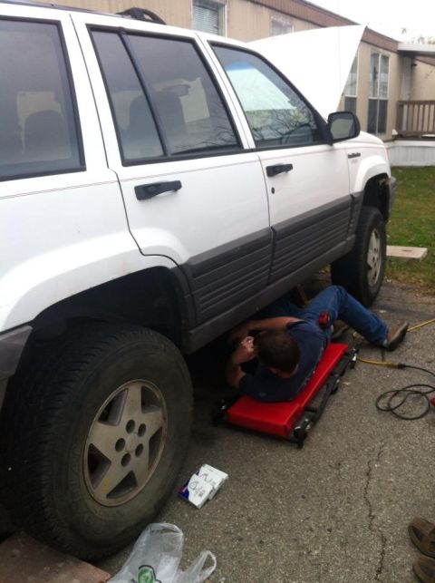 1989 Ford Bronco - Mechanic works on a white Jeep Grand Cherokee, hood up, on a driveway.