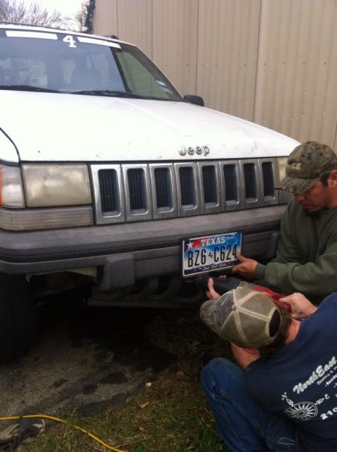 1989 Ford Bronco - Two men attach a Texas license plate to the front of a white Jeep Grand Cherokee.
