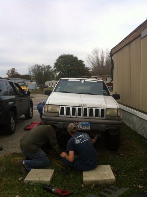 1989 Ford Bronco - Two people are working on the underside of a white Jeep Grand Cherokee, with another vehicle parked to its left.