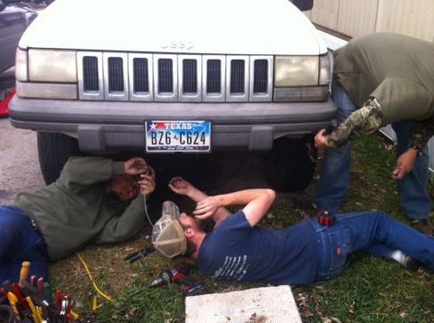 1989 Ford Bronco - Two men are working on the underside of a white Jeep Grand Cherokee, with a Texas license plate visible.