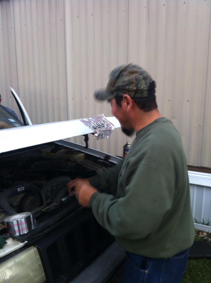 Man in camouflage hat works on car engine compartment; hood is open, showing engine parts and a roll of duct tape