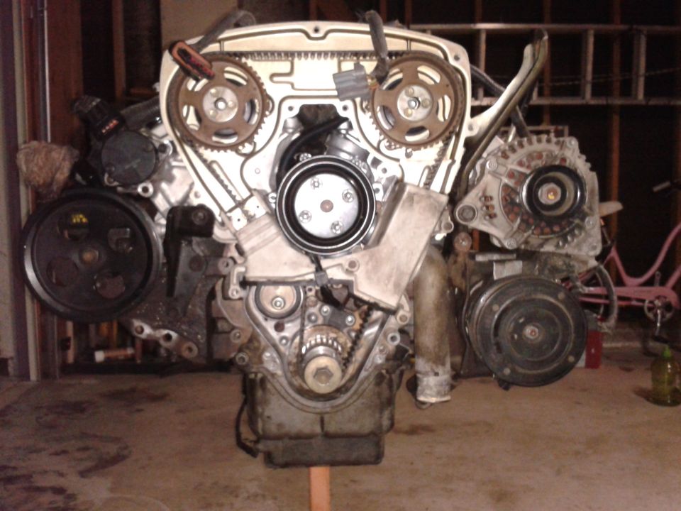 Engine assembly with timing belt, pulleys, alternator, and AC compressor visible. Mounted on a stand