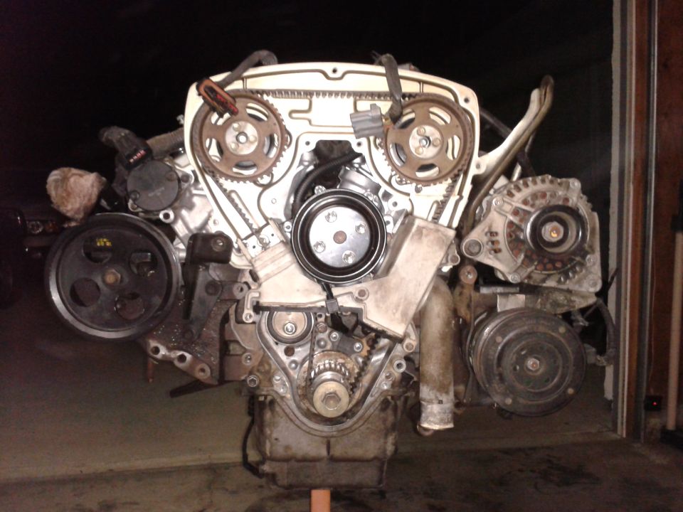 Engine assembly with timing belt, pulleys, alternator, and AC compressor visible. Mounted on a stand