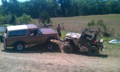 1987 Ford Bronco - A lifted Jeep tows a Ford Bronco through a grassy field with trees and a pond in the background.