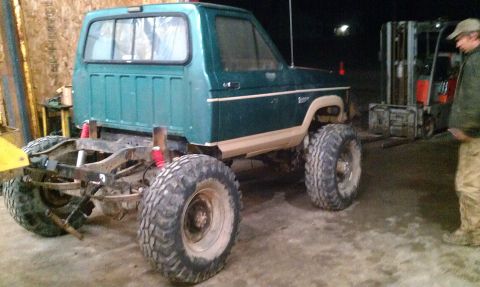 1987 Ford Bronco - A modified green pickup truck with oversized tires sits in a workshop, with a forklift visible in the background.