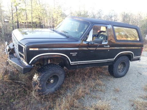1986 Ford Bronco - A black 1986 Ford Bronco is parked outdoors on a gravelly surface with dry grass and trees in the background.