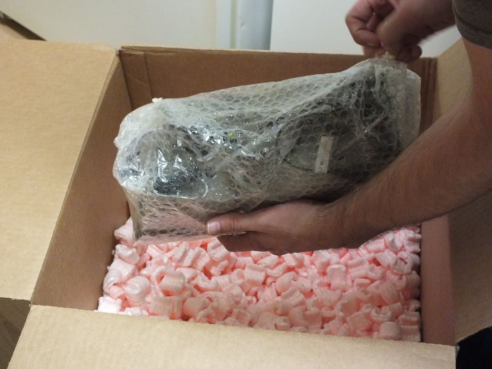 Hands holding a bubble-wrapped metal part above a box of pink packing peanuts