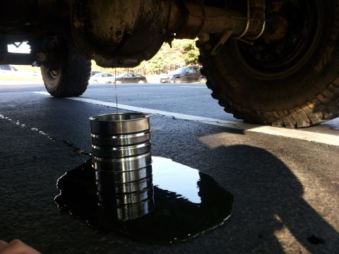 1986 Ford Bronco - Oil drains from a vehicle's undercarriage into a metal container on a paved surface.