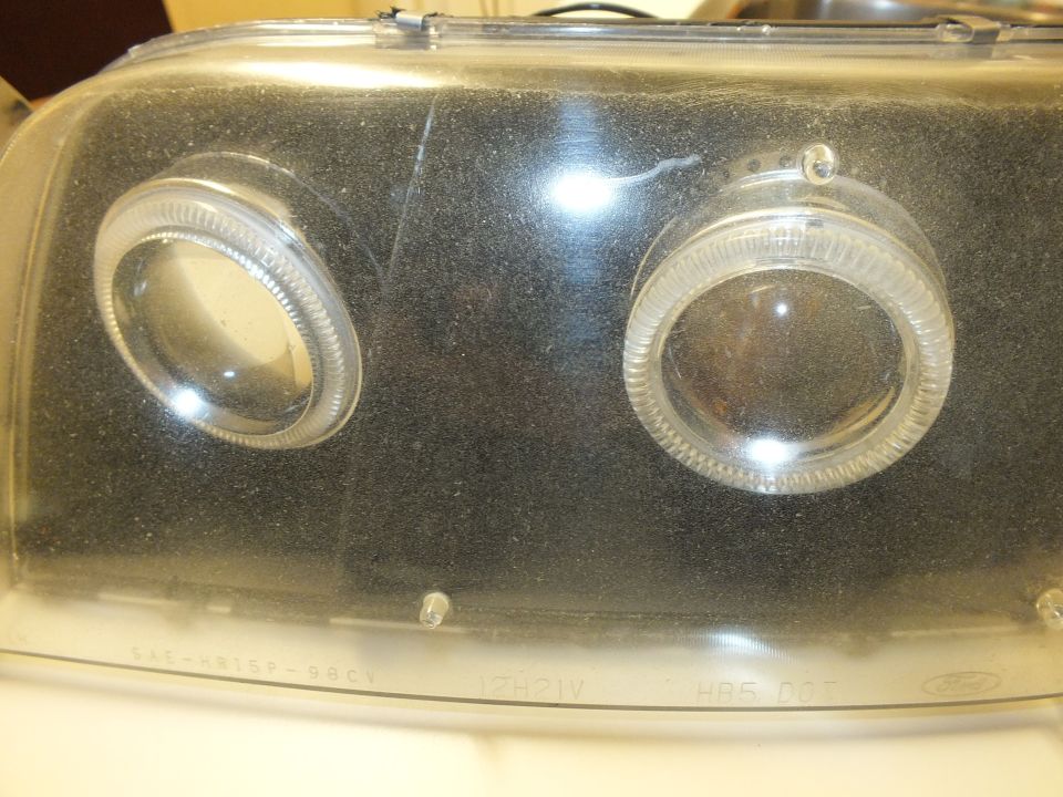 Close-up black projector headlight lens with halo rings and SAE 98CV text