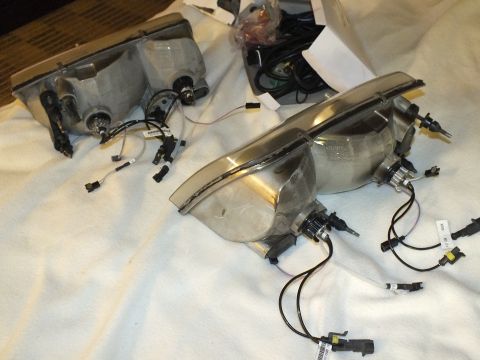 2003 Ford Crown Victoria - Two headlight assemblies in mid-build, one clear-lens unit with chrome trim and trailing connector harnesses.