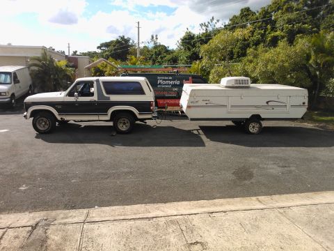1984 Ford Bronco - A white and brown 1984 Ford Bronco is seen from the side, towing a white pop-up camper.