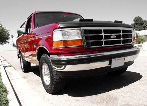 1994 Ford Bronco - Low passenger-side front three-quarter shot of a red full-size Ford Bronco 4x4 at a curb, edited so the truck stays in color while the back…