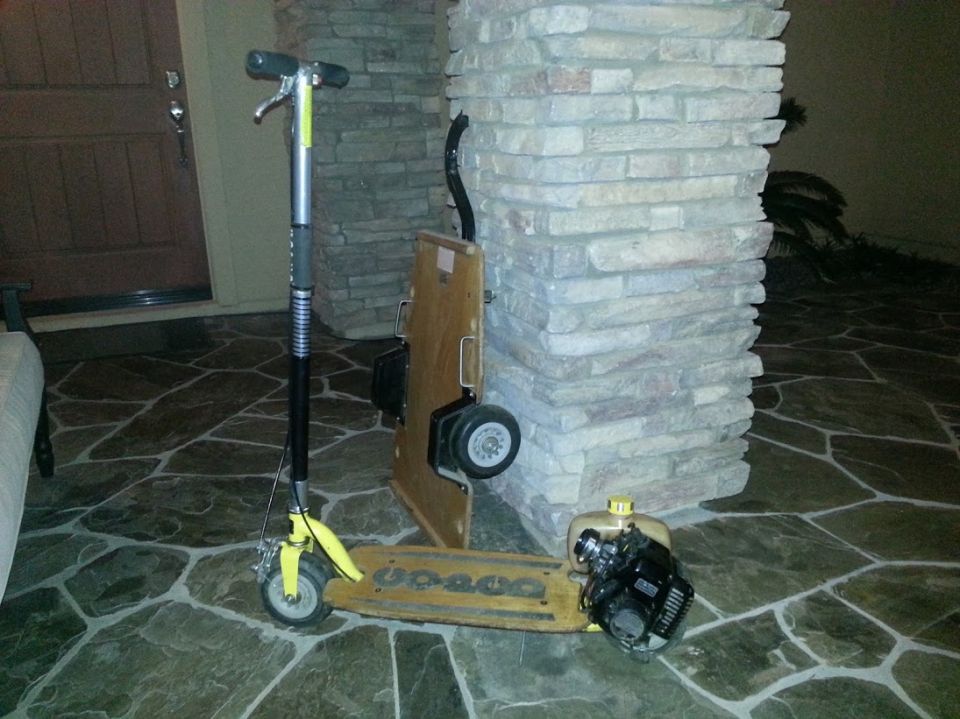 Yellow motorized scooter with a gas engine attached to the deck, leaning against a stone pillar