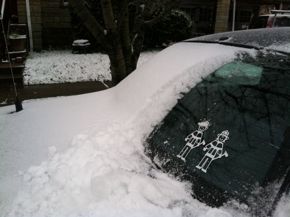 Snow-covered dark sedan rear window with two firefighter stick-figure decals in a snowy driveway