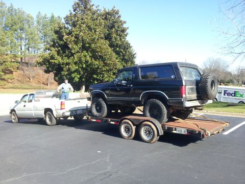 1986 Ford Bronco - A black 1986 Ford Bronco is loaded onto a trailer towed by a white pickup truck.