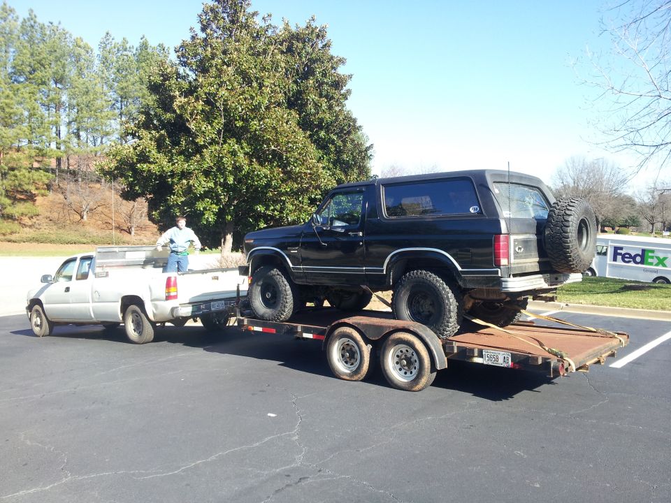 1986 Ford Bronco on a trailer being towed by a white pickup truck