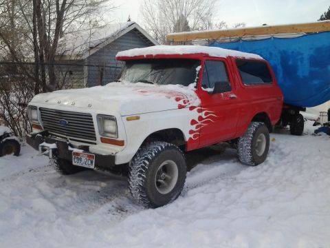 1979 Ford Bronco - A red and white Ford Bronco sits in the snow, partially covered by a blue tarp on a trailer.