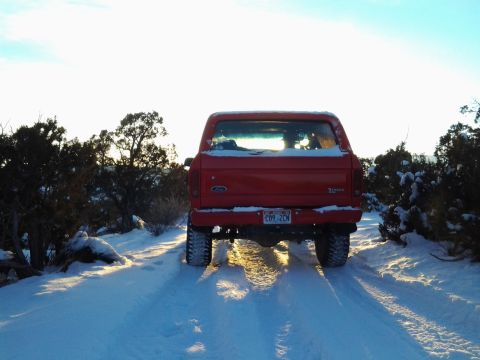 1979 Ford Bronco - A red 1979 Ford Bronco is seen from the rear, driving on a snow-covered road with sparse vegetation.