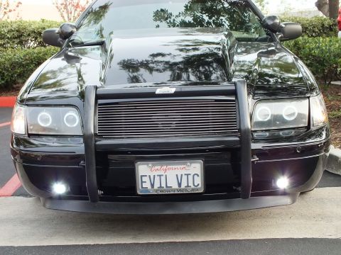 2003 Ford Crown Victoria - Tight shot of a black sedan front fascia, halo headlights, push bar and lit fog lamps over EVIL VIC plate.