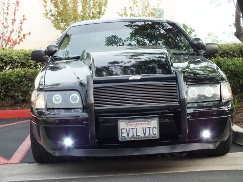 2003 Ford Crown Victoria - Black sedan straight on, mirrors out, EVIL VIC plate and lit fog lamps under a black push bumper.