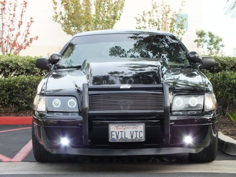 2003 Ford Crown Victoria - Head-on black Ford sedan, billet grille behind a grille guard, fog lights lit over EVIL VIC tag.