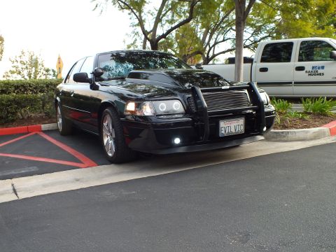 2003 Ford Crown Victoria - Angled black Ford sedan with grille guard and chrome wheels, white work truck parked alongside.
