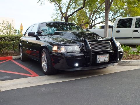 2003 Ford Crown Victoria - Side-front view of a black Ford sedan, EVIL VIC plate and grille guard, white pickup behind.