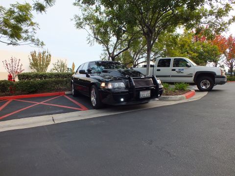 2003 Ford Crown Victoria - Wide lot shot of a black sedan with grille guard next to a white Chevy truck and shade trees.