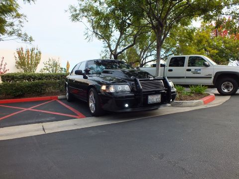 2003 Ford Crown Victoria - Distant front three-quarter of a black Ford sedan with push bar, white work truck alongside.