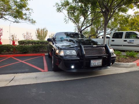 2003 Ford Crown Victoria - Angled black Ford sedan with grille guard and lit fog lamps, white pickup parked behind.