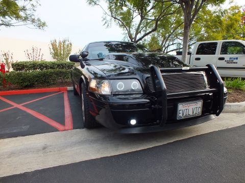 2003 Ford Crown Victoria - Glossy black sedan angled in a lot, EVIL VIC plate and billet grille, work truck alongside.