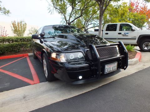 2003 Ford Crown Victoria - Angled front of a black Ford sedan, EVIL VIC plate and chrome wheels, trees and pickup behind.