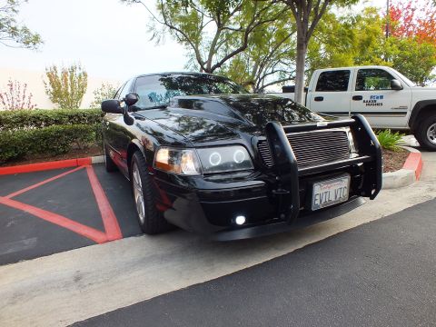 2003 Ford Crown Victoria - Black Ford sedan angled in lot, billet grille and chrome wheels, white work truck in background.