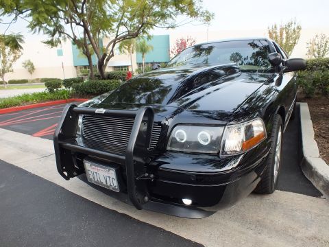 2003 Ford Crown Victoria - Angled front of a glossy black Ford sedan with grille guard and chrome wheels at a lot.