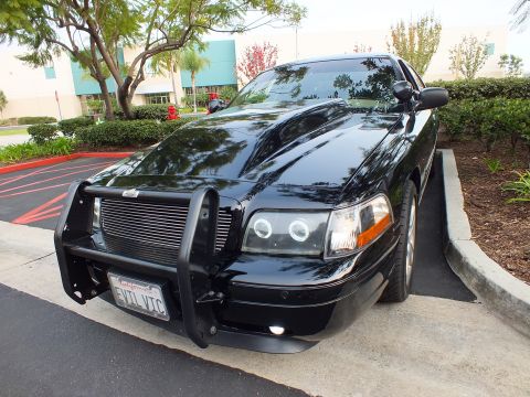 2003 Ford Crown Victoria - Black Ford sedan angled in a lot, billet grille and EVIL VIC plate, building and trees behind.