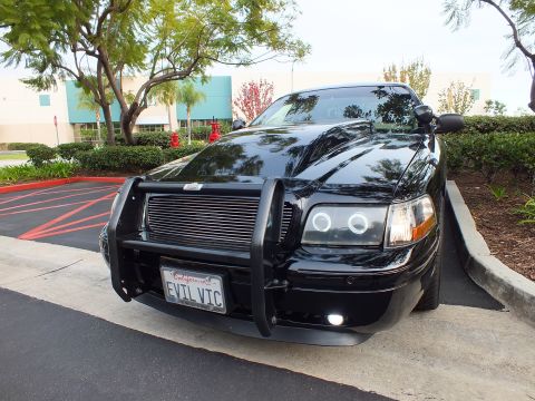2003 Ford Crown Victoria - Angled front of a black Ford sedan with grille guard and lit fog lamp, parked by a building.